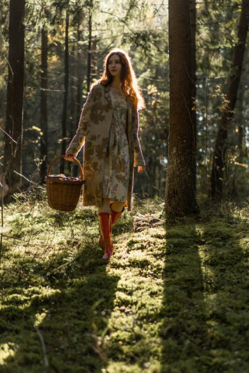 Alone in the forest, a young woman with flowing red hair and a linen dress carefully gathers mushrooms