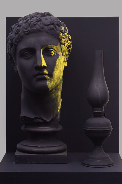 A lume spento, 1985 plaster cast, lamp 54 x 41 x 32 cms. In Parmiggiani's striking composition, a golden glow illuminates a fragment of antiquity and a lamp, both rendered in stark relief against darkness. It speaks to the fading of old ideals, or perhaps a singular, enduring truth against a backdrop of oblivion. Image courtesy of the Artist/Estorick Collection of Modern Italian Art