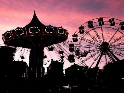 A photo showing the silhouettes of a Ferris wheel and a carousel at dusk, glowing against a pink evening sky at the fair