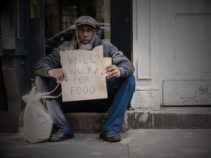 A mature man sits in a doorway holding a cardboard sign, representing homelessness and poverty in the United States