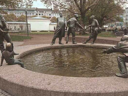 La fuente está rodeada irregularmente por un conjunto de esculturas de bronce distribuidas alrededor de la pileta de hormigón. El agua gira en sentido contrario a las agujas del reloj, en la dirección del flujo de dinero. Karl-Henning Seemann,1976. "Ciclo del Dinero" Aquistrán, Alemania