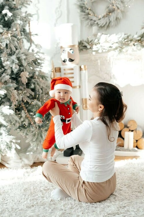 A little baby dressed in a Santa costume sits on his mother's lap, nestled beside the twinkling Christmas tree