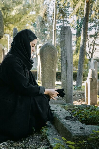 In a cemetery, a woman dressed in black and wearing a hijab sorrows for the dead, praying in accordance with Islamic tradition