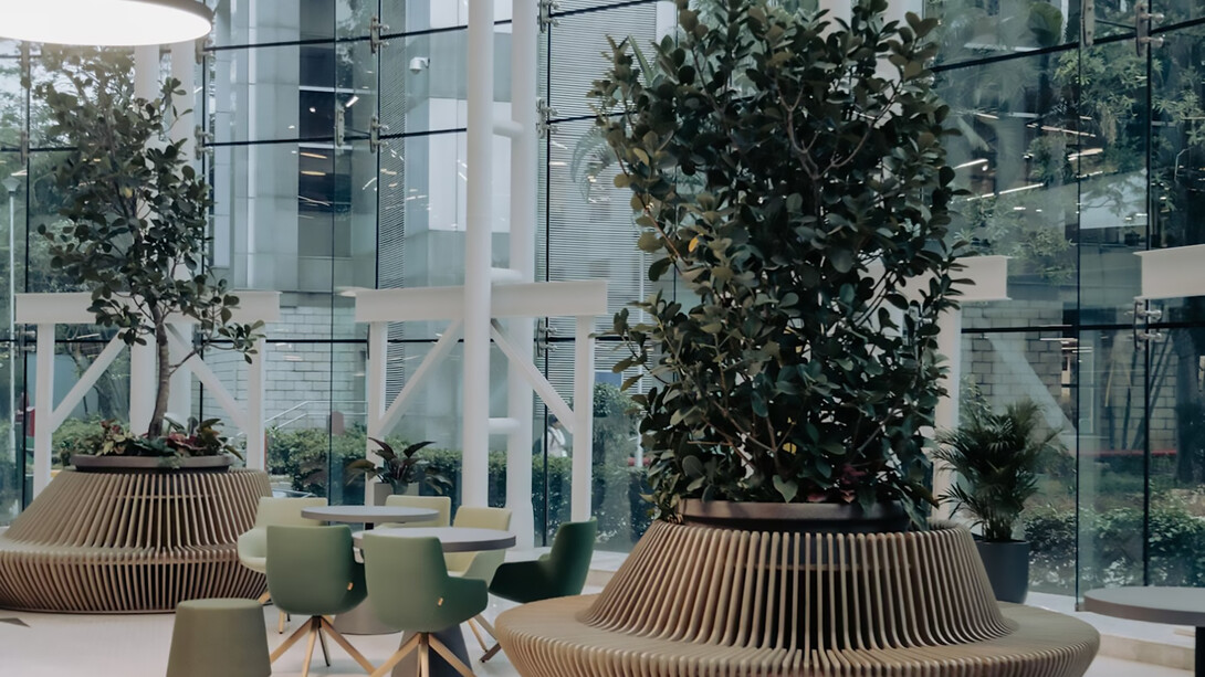 Modern office lobby with floor-to-ceiling windows, green chairs around round tables, and ribbed planters holding lush indoor plants. Bright and airy with a contemporary feel