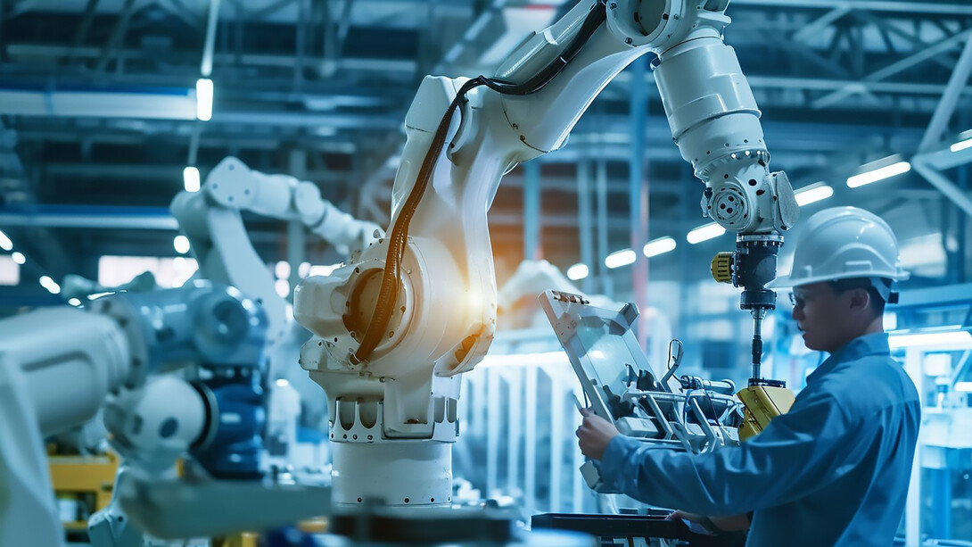 In a brightly lit, advanced manufacturing facility, a worker wearing a safety helmet closely monitors a state-of-the-art robotic arm in operation, ensuring precision and safety on the production line
