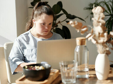 A girl with Down syndrome uses a laptop while sitting on a couch, highlighting the role of disability technology in everyday life