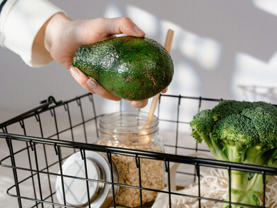 A person holding a green avocado above a black steel basket, representing antioxidant foods, iron-rich plant foods, healthy superfoods, and natural supplements.