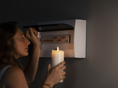 Una donna con una candela accesa in mano osserva un quadro elettrico aperto sulla parete, durante un blackout