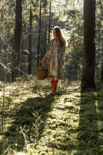 A young woman with long red hair, wearing a linen dress, wanders alone in the forest while collecting mushrooms