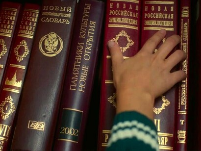 Pessoa seleciona livro de enciclopédia em uma prateleira. Louis foi se empolgando aos poucos e contou os detalhes que achou necessários, sobre a entrevista com Eugène Hyndormais e seu projeto de um mundo a partir da influência cultural do Egito, e não de Grécia e Roma