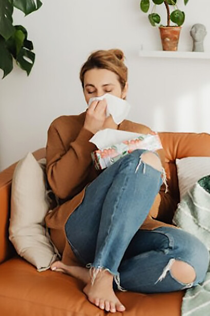 Woman seated on a sofa, using a tissue to blow her nose due to allergy symptoms