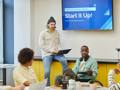 A man presents in front of his startup team in a meeting room, guiding a brainstorming session where founders and colleagues collaborate on their business pitch