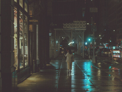 Rain falls on a city street at night, neon lights reflecting off wet asphalt in a noir-style cityscap