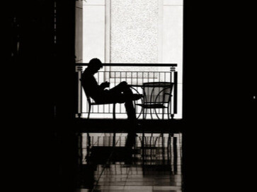 Assis dans la pénombre, il dialogue avec un écran. Le visage effacé, la présence réduite à un signal. Le lien humain, lui, s’efface dans la lumière blanche des pixels