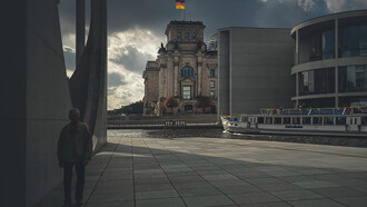 An elderly man walks along a gray concrete path beside a brown building in Berlin, Germany