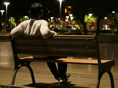 A lone man on a bench in a nighttime city park evokes themes of isolation, urban loneliness, and a society that feels increasingly disconnected