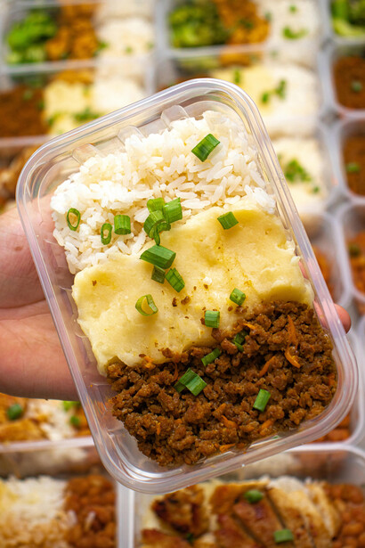 A hand holding a meal prep container filled with rice, mashed potatoes, and meat