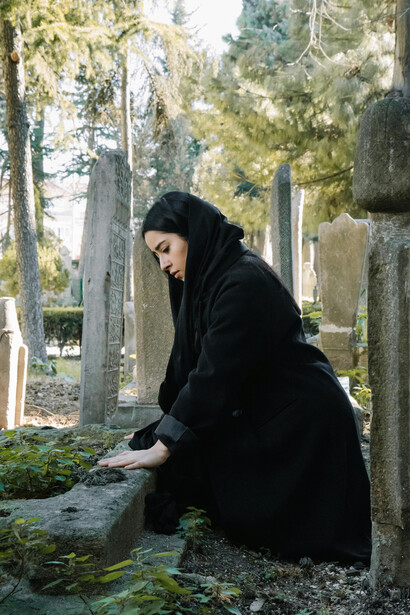 A grieving woman in a black hijab prays at a gravesite, reflecting Islamic mourning practices