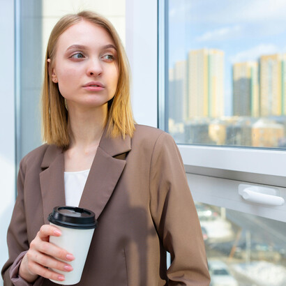 A woman in a brown blazer holds a cup of coffee, lost in thought