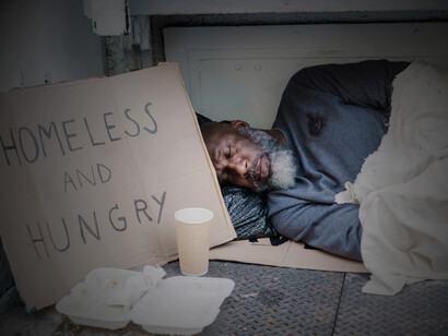 A photo of a homeless man sleeping on the ground with a cardboard sign placed beside him, in the US
