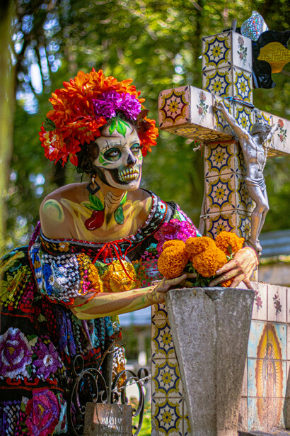 Mexico City, Mexico. A vibrant Day of the Dead Catrina in a cemetery with a Día de los Muertos altar and a woman wearing colorful makeup