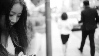 A woman uses her smartphone on the sidewalk while a man and woman walk away from her