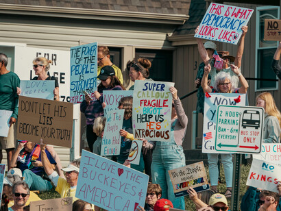 A diverse group of citizens holding protest signs in a suburban street, representing the awakening of democratic agency in the face of institutional fatigue