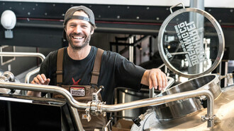 A smiling man standing near metal brewing tanks, embodying the passion behind craft beer production