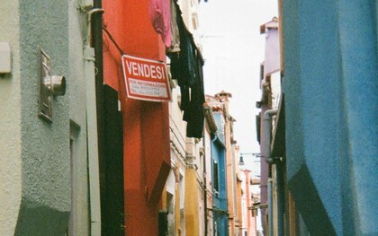 Un vicolo stretto a Burano, Italia, fiancheggiato da edifici dai colori vivaci, con un cartello rosso con la scritta "Vendesi" su uno degli edifici