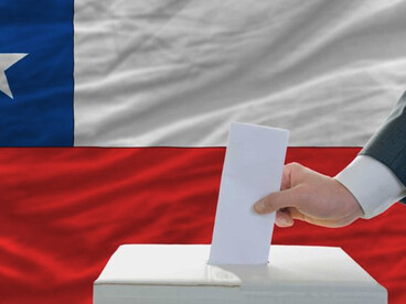 Chile celebró recientemente la primera vuelta de sus elecciones generales el 16 de noviembre de 2025, para elegir un nuevo presidente, todos los miembros de la Cámara de Diputados y la mitad de los escaños del Senado. Una mano depositando su voto en una urna, con la bandera nacional de Chile de fondo