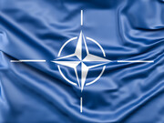 The NATO symbol resting on blue satin, conveying how global order is often staged as harmony while driven by strategic profit