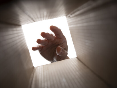 Una mano vista dall'interno di una scatola di cartone, che scende verso il fondo. L'effetto del raggio di luce che si diffonde dall'apertura aggiunge un senso di mistero