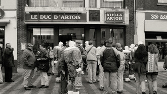 A black-and-white photograph of people standing in a queue outside a shop, Belgium