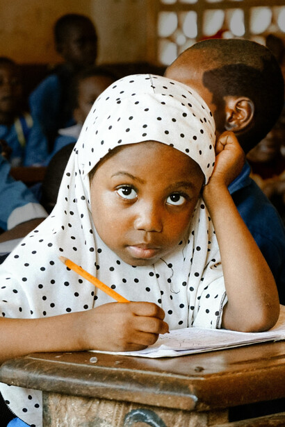 A young girl in Africa focuses on her writing during class, surrounded by classmates, capturing the spirit of learning and academic growth