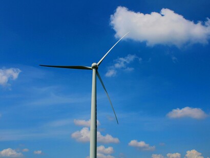 A single wind turbine with three blades stands tall against a clear blue sky with scattered white clouds, representing the increasing adoption of wind energy as a key renewable resource