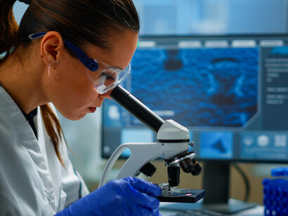 Primo piano di una scienziata con guanti e occhiali protettivi che guarda attraverso un microscopio in un laboratorio di ricerca medica, con un monitor che mostra un'immagine biologica sfocata sullo sfondo