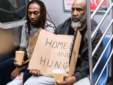 Two people seated in a subway, holding a simple sign that says they are homeless, symbolising how lives are disrupted by eviction