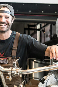 A smiling man standing near metal brewing tanks, embodying the passion behind craft beer production