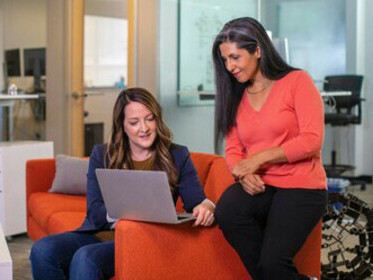 Two businesswomen discussing sales while sitting on a sofa, with a laptop open in front of them