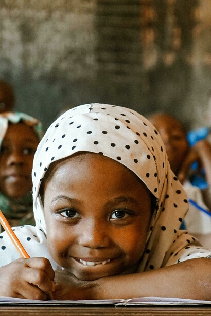 An African schoolgirl smiles while writing with a pencil in class, as fellow students study in the background, symbolizing academic pursuit and childhood education