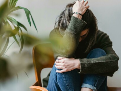 Una donna seduta su una sedia, con le ginocchia raccolte al petto, affonda la testa tra le braccia. La sua postura chiusa e il volto nascosto suggeriscono profonda tristezza, ansia o disperazione