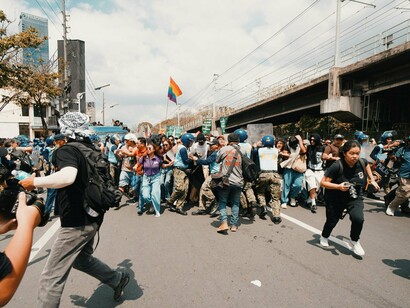 Protesters pushing through police defences amid a noisy demonstration, reflecting the clash between youth activism and power structures