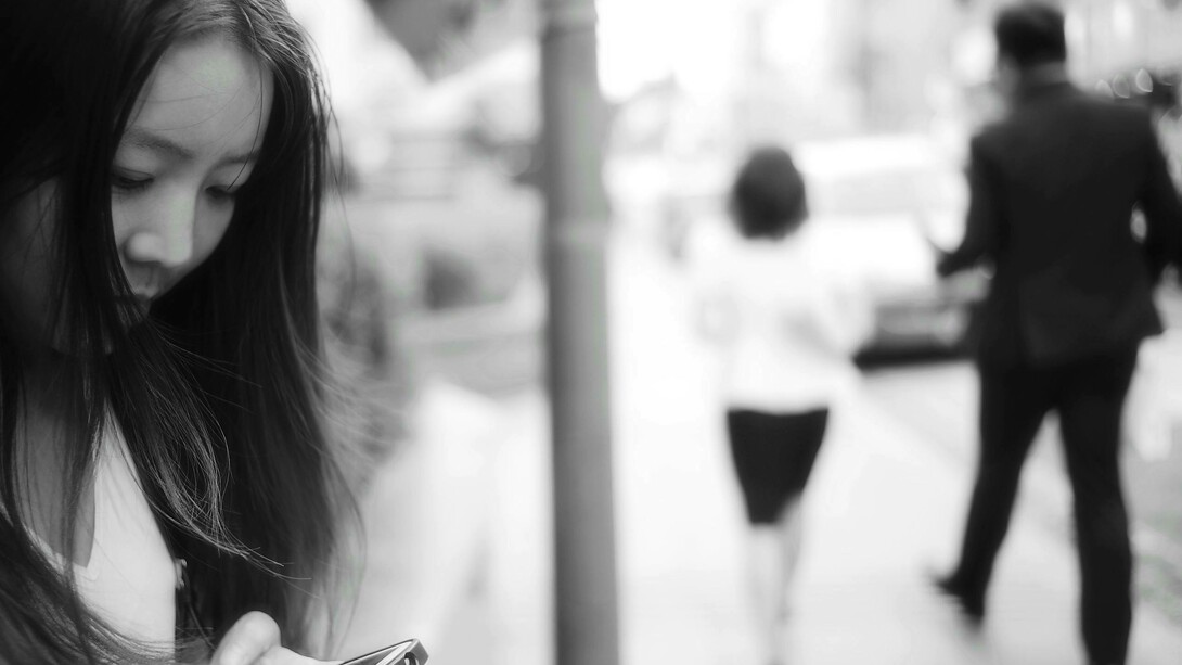A woman uses her smartphone on the sidewalk while a man and woman walk away from her