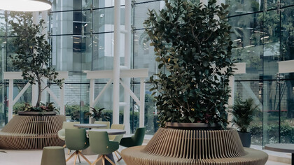 Modern office lobby with floor-to-ceiling windows, green chairs around round tables, and ribbed planters holding lush indoor plants. Bright and airy with a contemporary feel