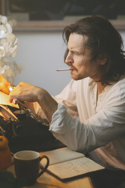 In a dimly lit room, a solitary poet sits at his desk, cigarette in hand, lost in thought over a sheet of paper