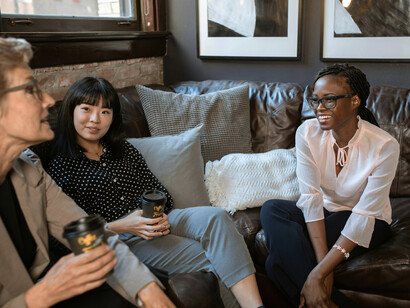 A Black woman, an Asian woman, and a Caucasian woman holding disposable cups and having a conversation at work