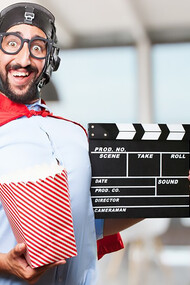 A man dressed in Superhero attire holds popcorn in one hand and a black movie production clapper board 