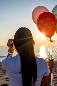 A woman standing on the beach, holding colorful balloons, with her back to the camera