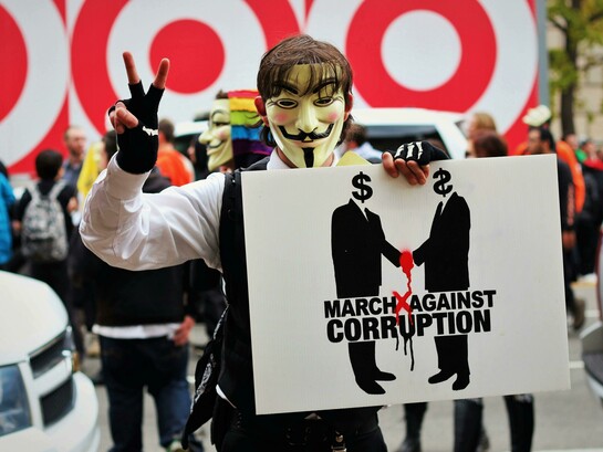 Person wearing a Guy Fawkes mask and holding a sign that reads “March Against Corruption”