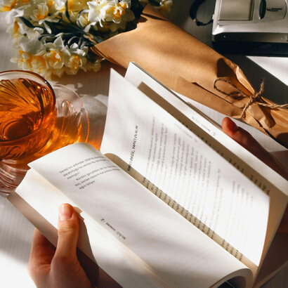 A woman’s hands hold a book, with a cup of tea and a bouquet of daffodils placed beside her